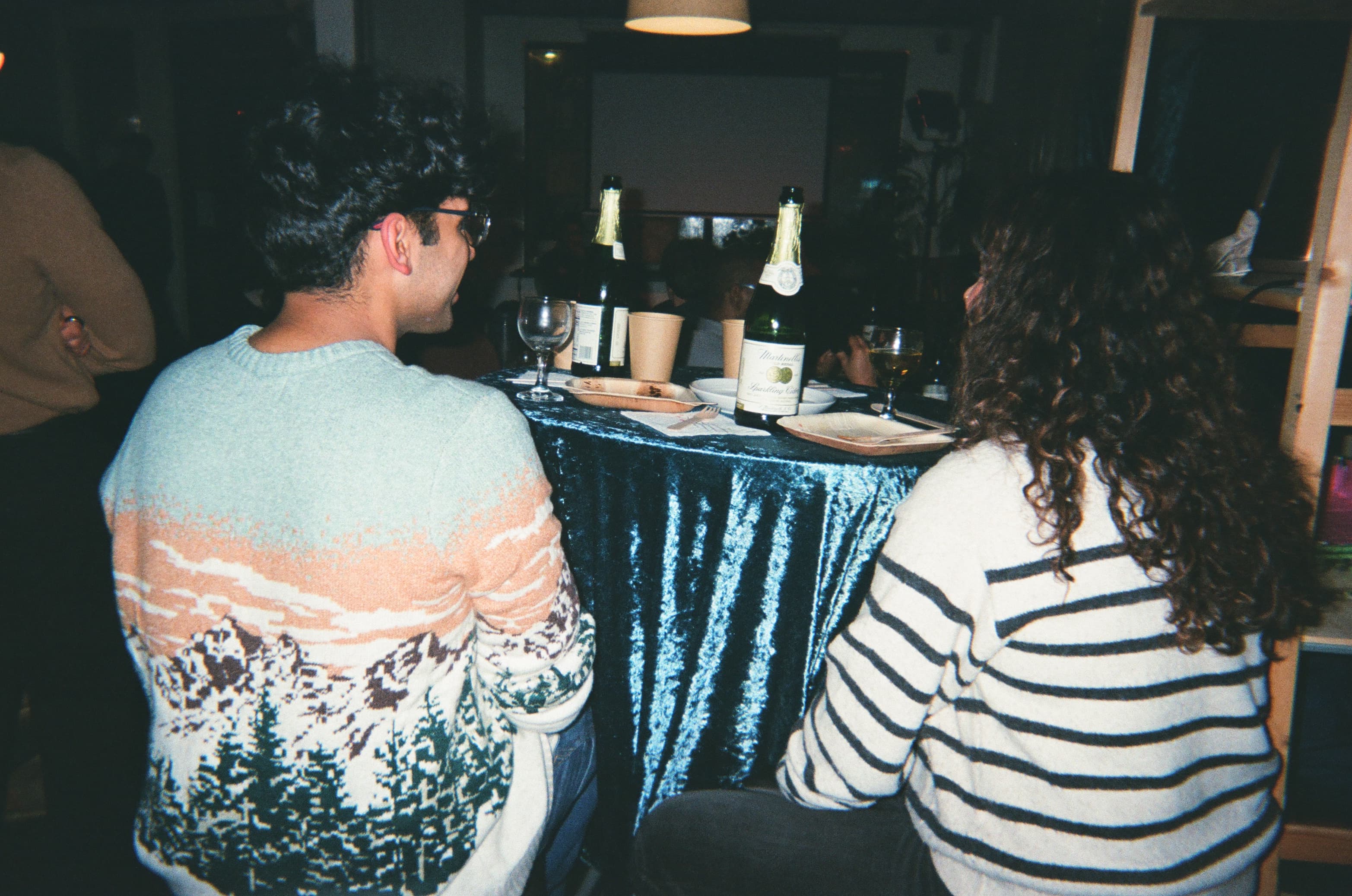 Two members in conversation at a dinner table covered in blue velvet, lit by a single warm pendant. Boston, MMXXVI.