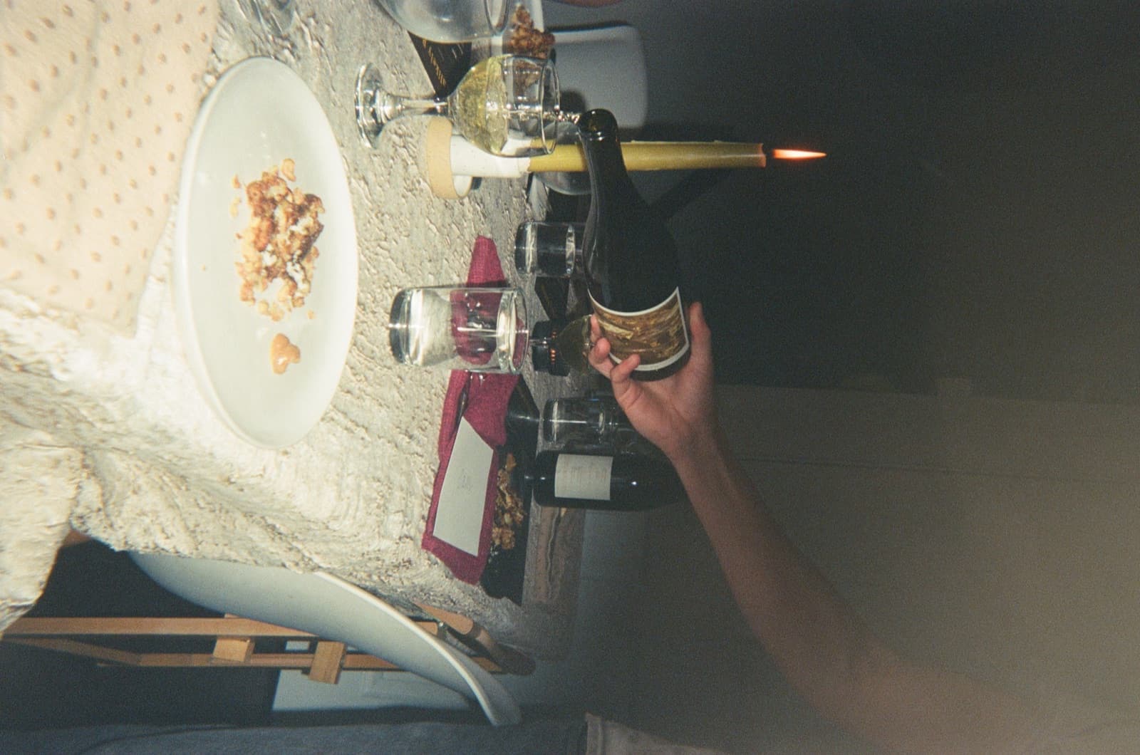 A hand pouring wine on a cream tablecloth; a candle in the foreground.